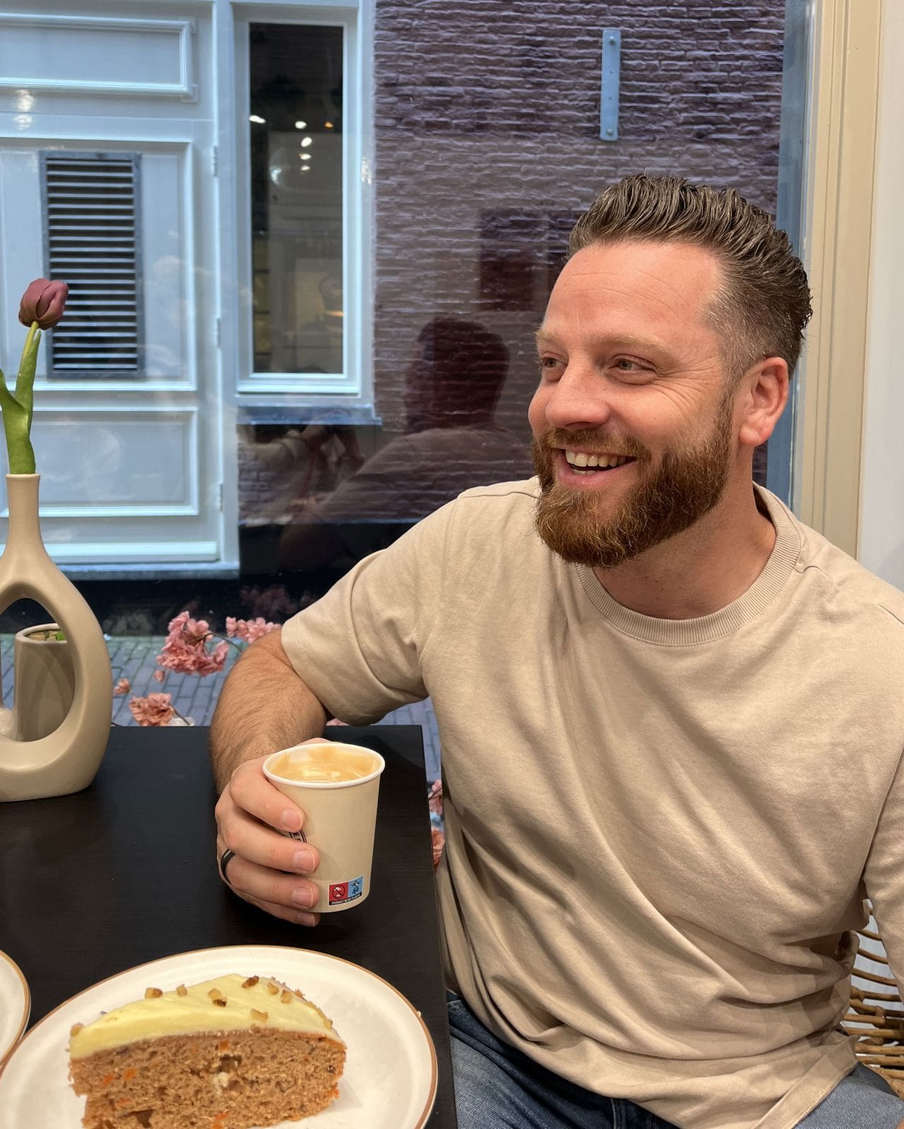 Roel Willemse lacht terwijl hij koffie drinkt in een café, met een stuk worteltaart op tafel.