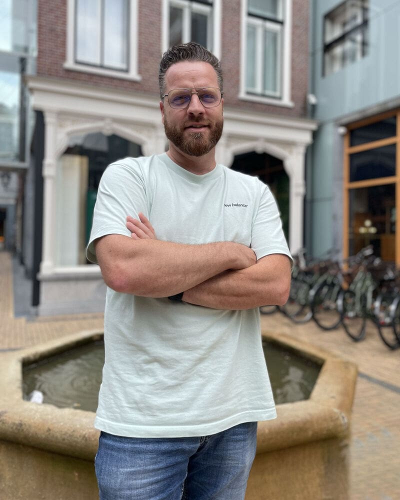 Roel Willemse staat met gekruiste armen bij een fontein in een stedelijke omgeving, in een casual outfit